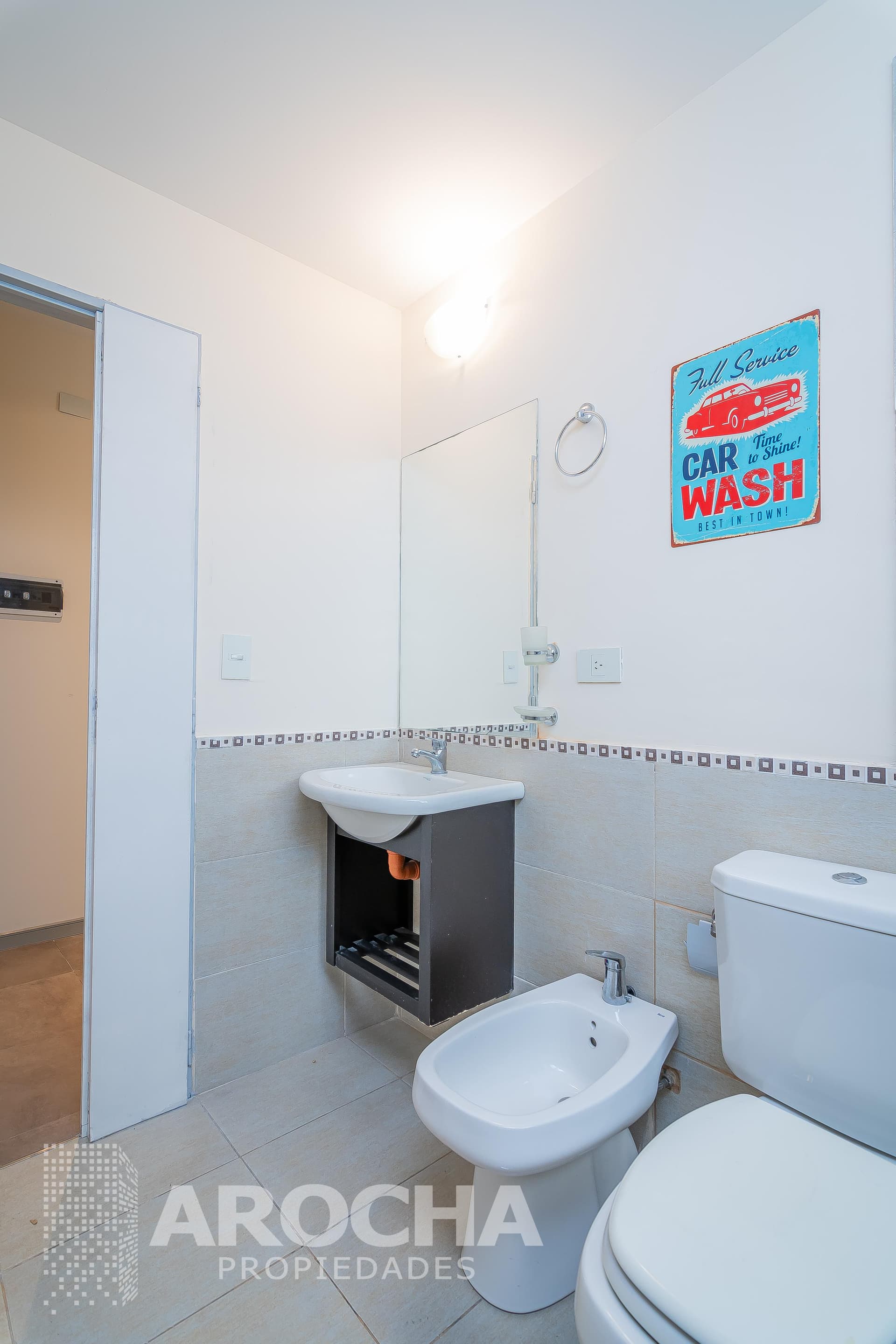 Baño completo con inodoro, bidet, lavatorio, espejo y puerta de entrada.
