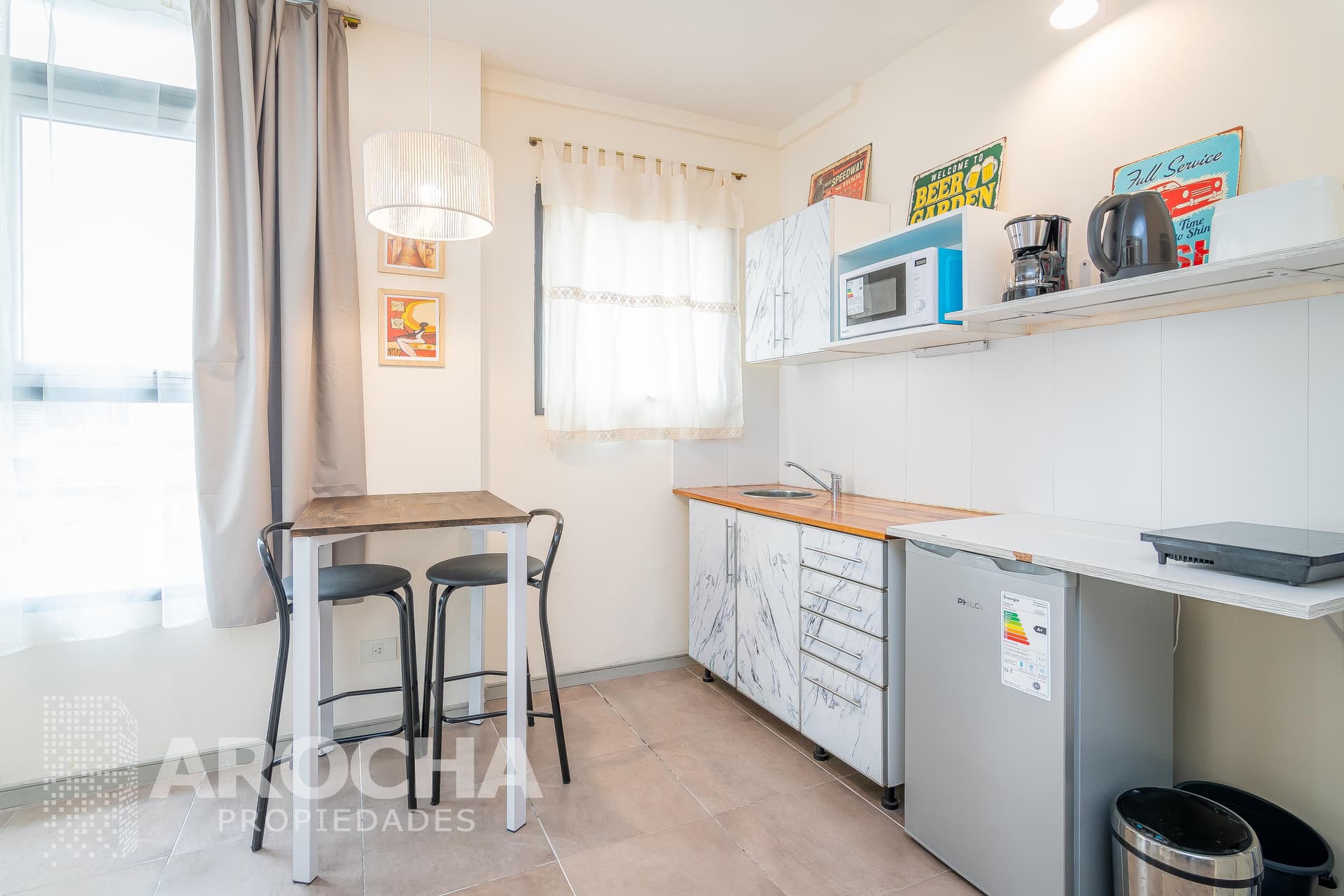 Cocina con bacha, heladera, microondas, anafe eléctrico y mesa alta con banquetas.