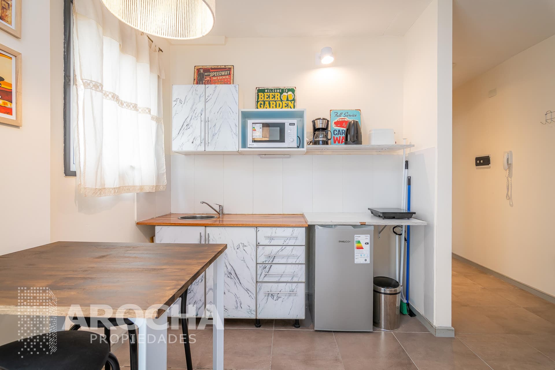 Cocina con alacenas blancas, bacha, heladera, mesa y vista al pasillo.