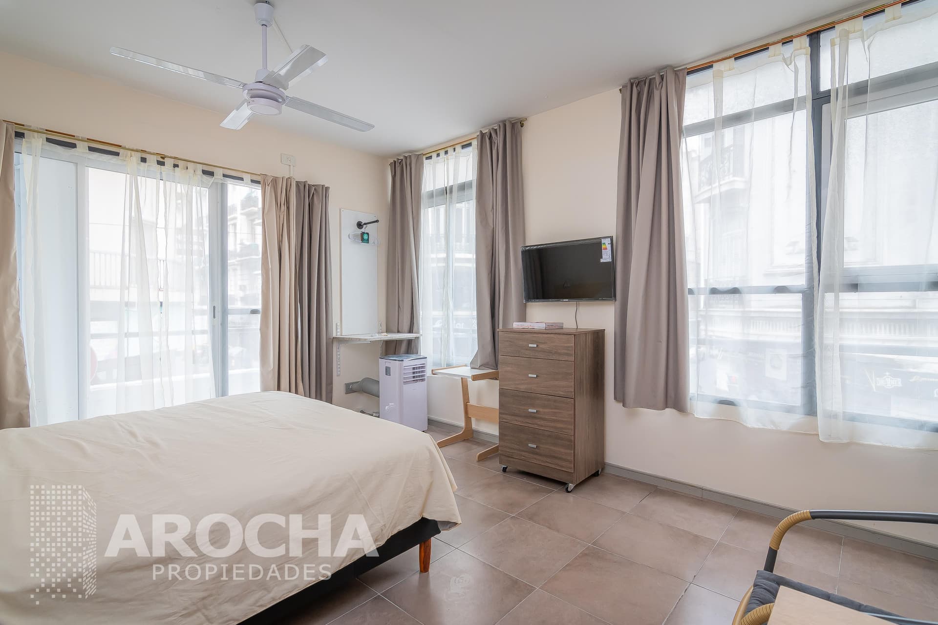 Dormitorio con cama, grandes ventanales, televisor en pared y cómoda de madera.