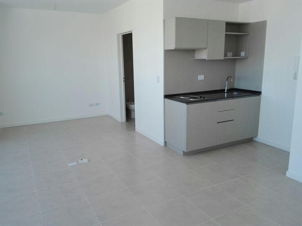 Living vacío con cocina integrada y puerta que conduce al baño.