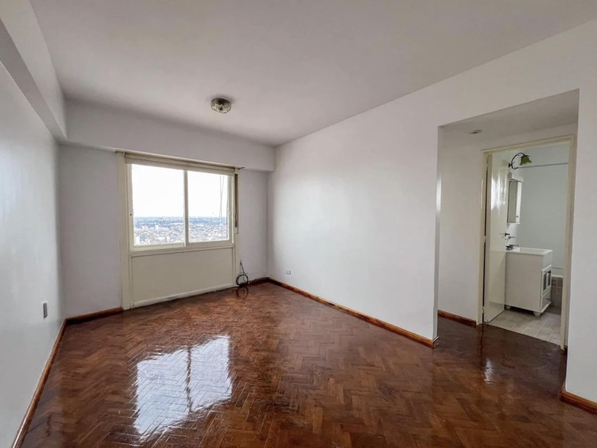 Living vacío con piso de parquet, ventana y acceso al baño