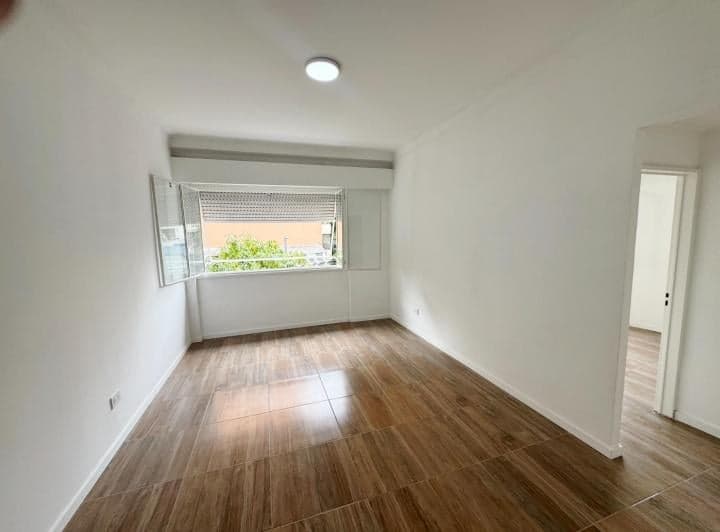 Dormitorio vacío con ventana y persiana, pisos cerámicos y paredes blancas.