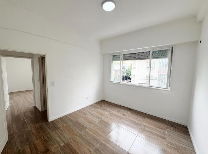 Dormitorio vacío con ventana, persiana, pisos cerámicos y acceso a otra habitación.