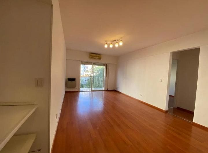 Living vacío con piso de madera, paredes blancas y puerta que da a un balcón.