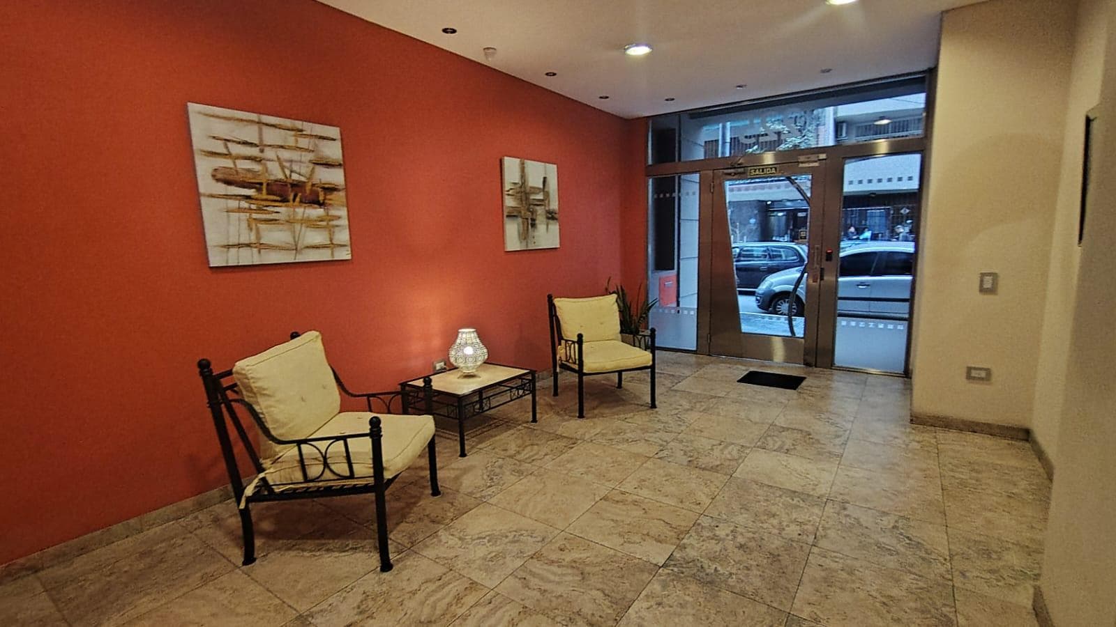 Hall de entrada del edificio con sillones, pared roja y puerta de vidrio.