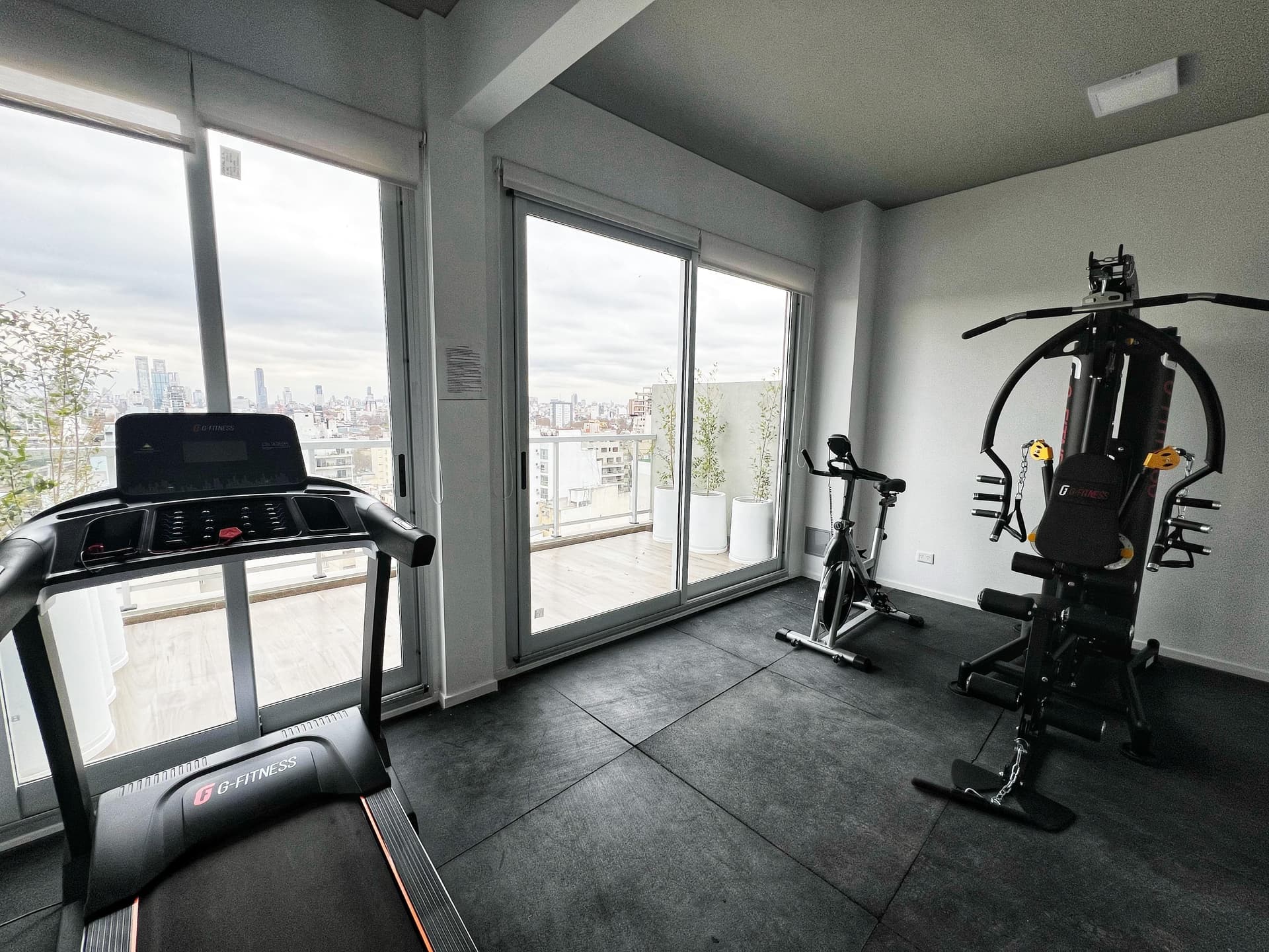 Gimnasio del edificio con cinta de correr y bicicleta fija frente a grandes ventanales.