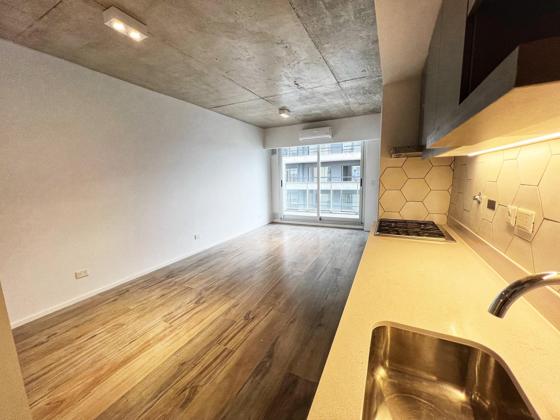 Living del departamento con cocina integrada, piso de madera y vista al balcón.