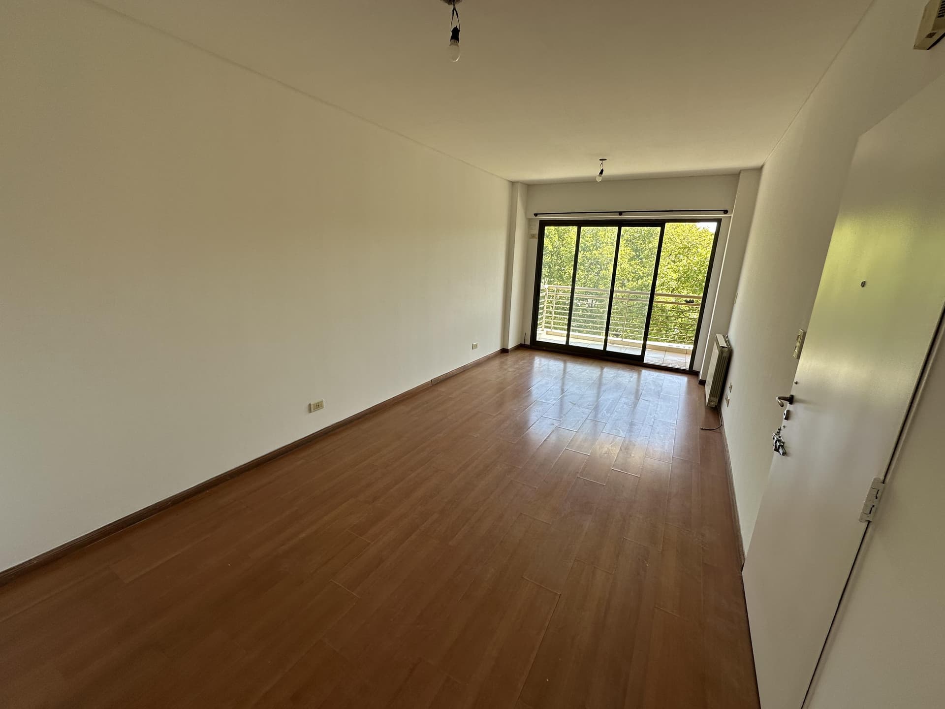 Living vacío con piso de madera y salida a balcón.