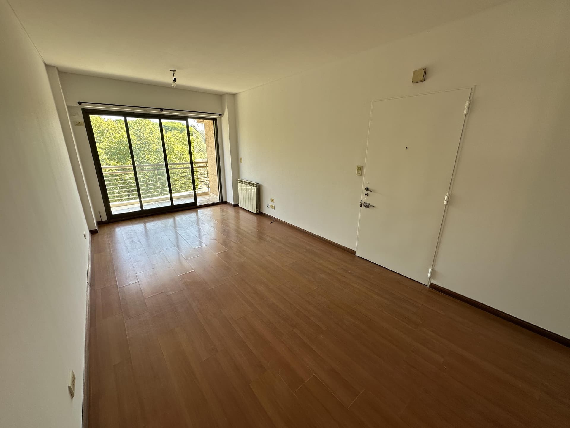 Living vacío con piso de madera, ventana grande y puerta de entrada.