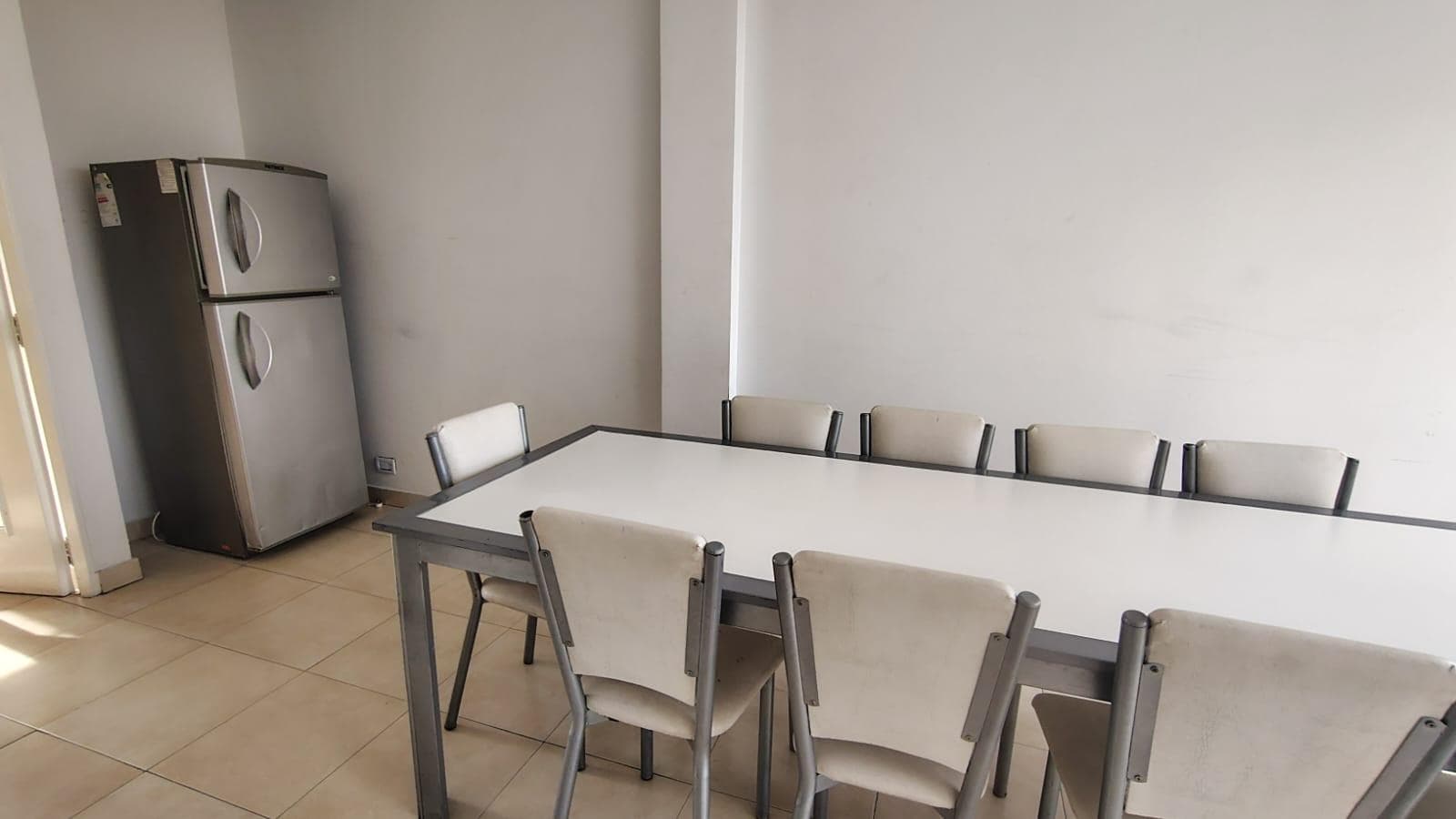 SUM con mesa larga y sillas, heladera, piso de baldosas.