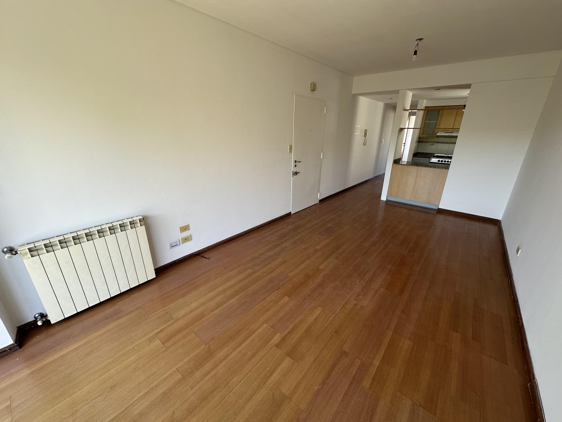 Living vacío con piso de madera, radiador y conexión a cocina.