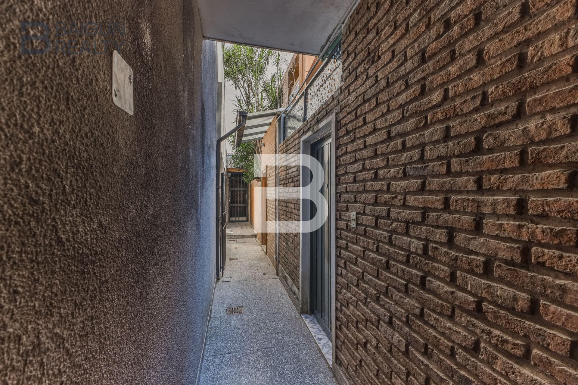 Pasillo exterior angosto con paredes de ladrillo y rejas.