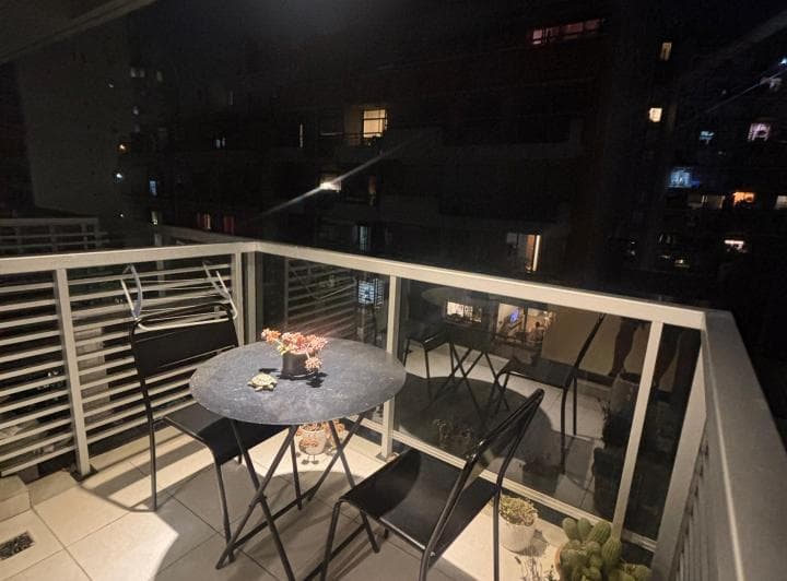 Balcón de noche con mesa y sillas de exterior, con edificios iluminados.