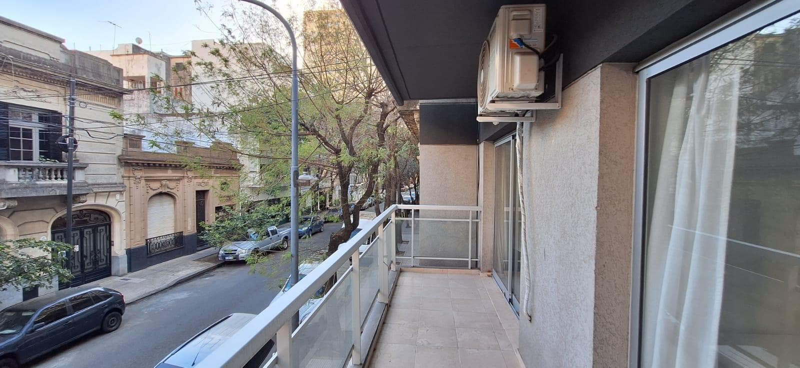 Balcón angosto con baranda de vidrio, aire acondicionado y vista a la calle.