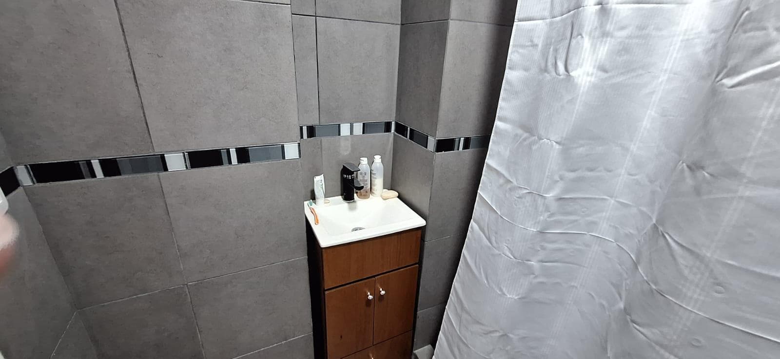 Baño con azulejos grises, lavatorio pequeño y cortina de ducha.
