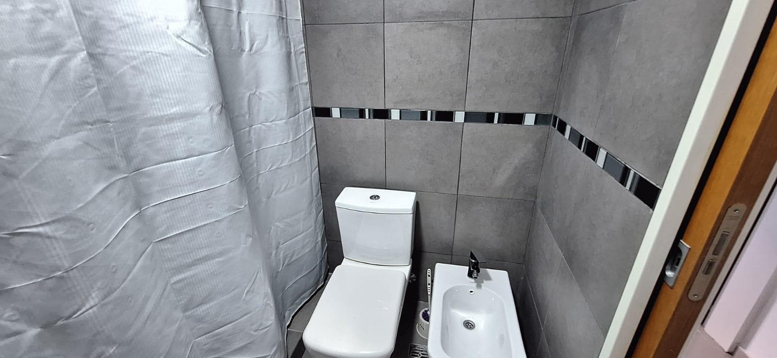 Baño con azulejos grises, inodoro, bidet y cortina de ducha.
