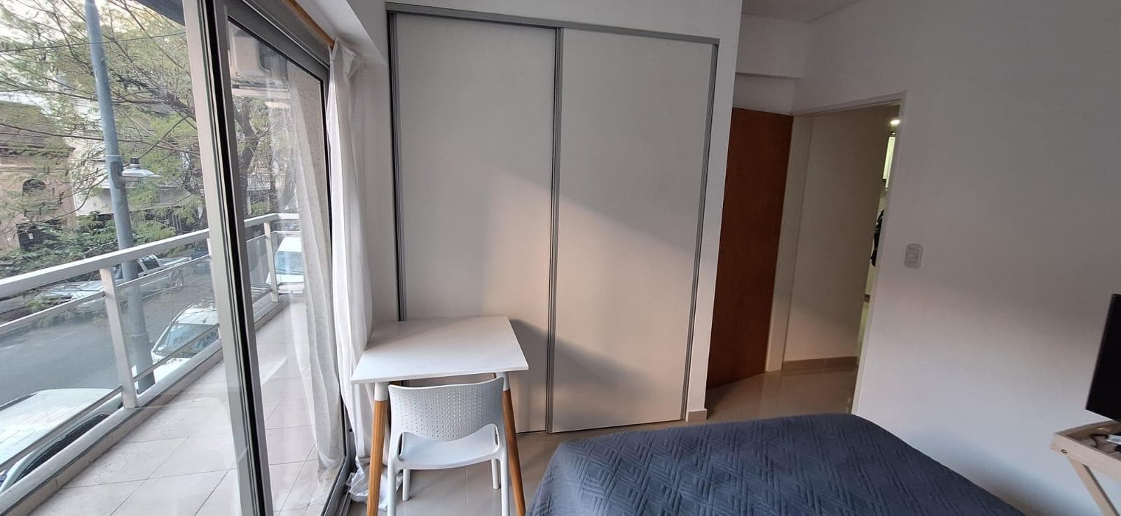 Dormitorio con cama, placard blanco y escritorio junto al balcón.