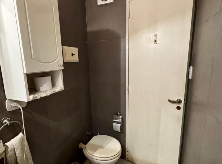Baño con inodoro y mueble de guardado superior, paredes de azulejos grises.