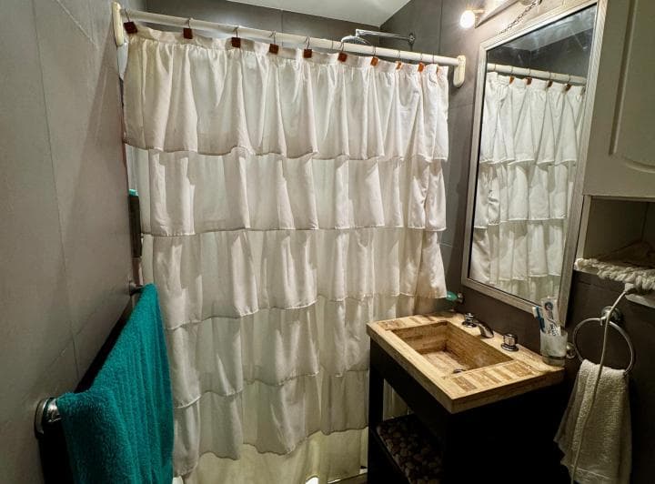 Baño con ducha con cortina blanca, vanitory de madera y espejo.