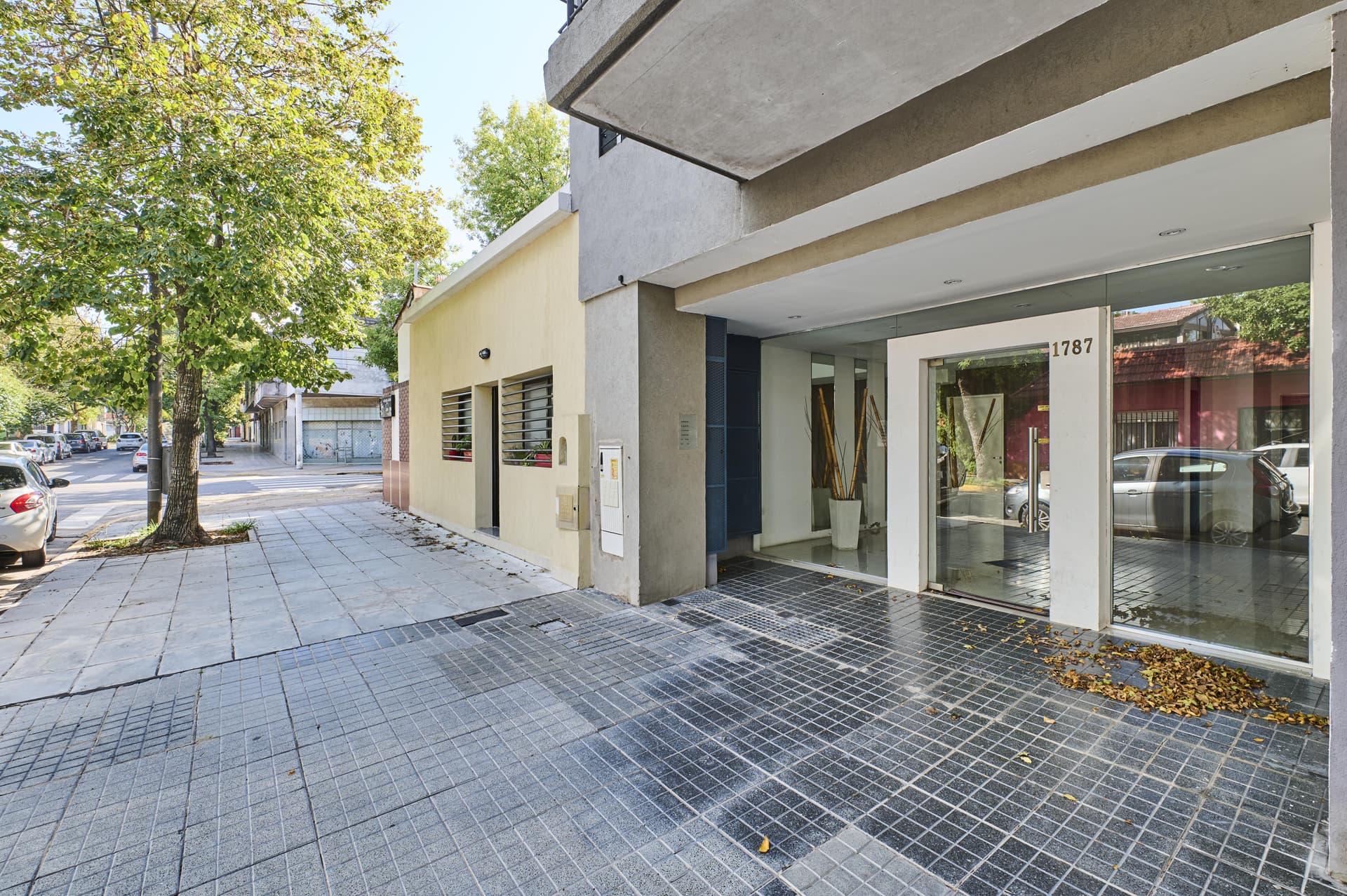 Fachada del edificio con entrada de vidrio, árboles y vereda.