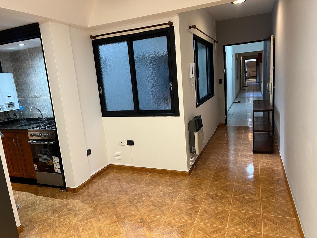 Living vacío con ventanas y estufa, conectando a un pasillo y cocina.