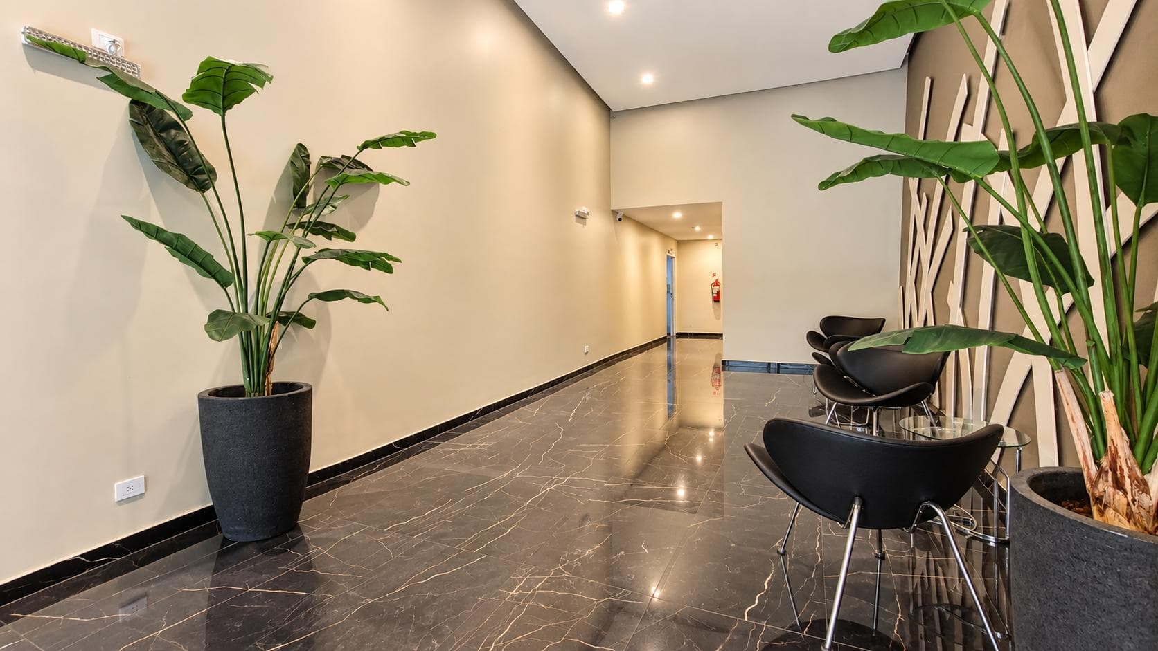 Hall de entrada del edificio con sillones negros, plantas grandes y piso oscuro.