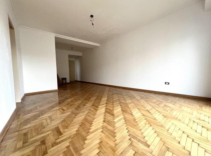 Living vacío con piso de madera, paredes blancas y vista a un pasillo.
