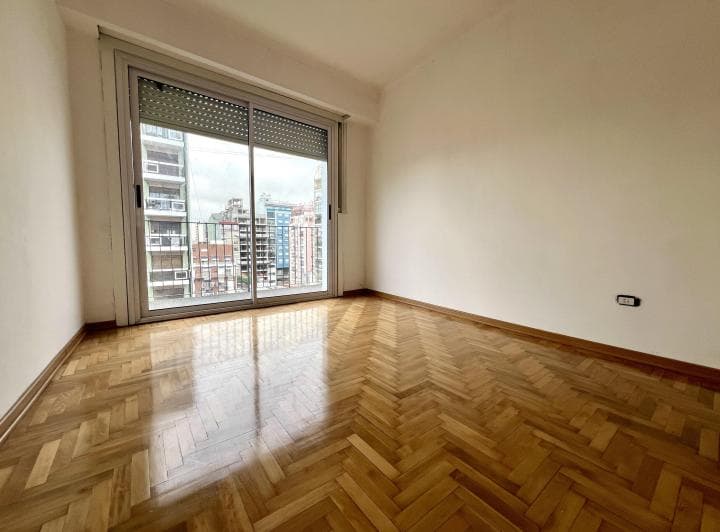 Dormitorio vacío con piso de madera y gran ventanal con persiana.