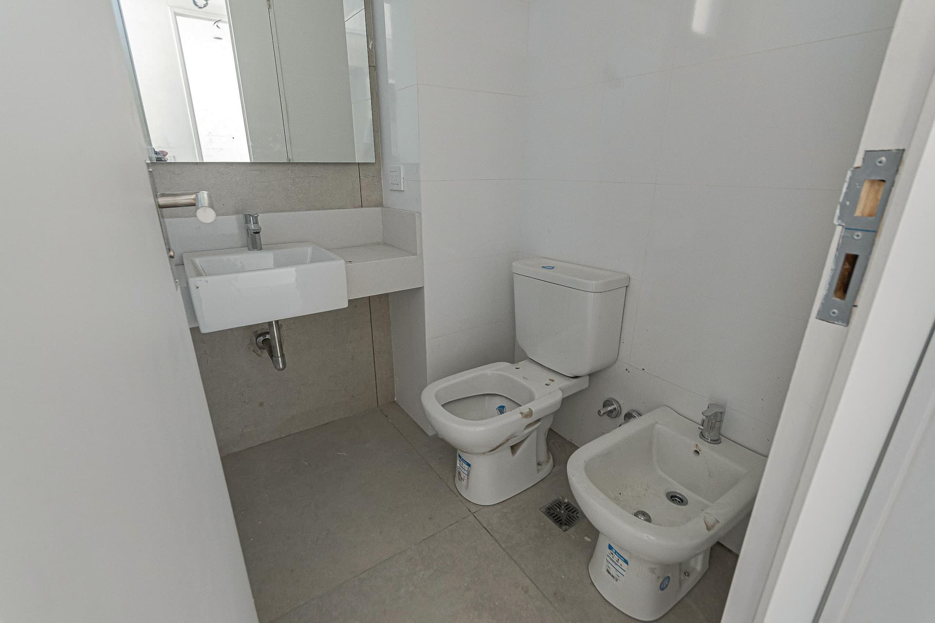 Baño con inodoro, bidet, lavatorio con espejo y paredes blancas.