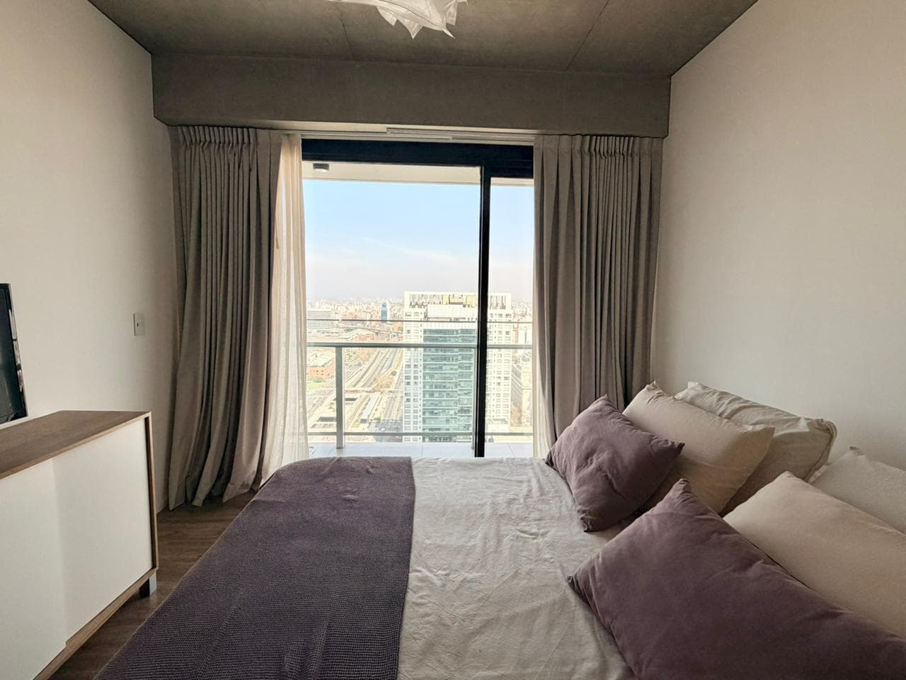 Dormitorio con cama doble, cortinas grises y ventanal con vista a la ciudad.