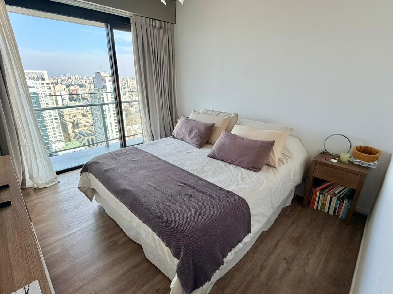 Dormitorio con cama doble, mesa de luz y ventanal con vista urbana.