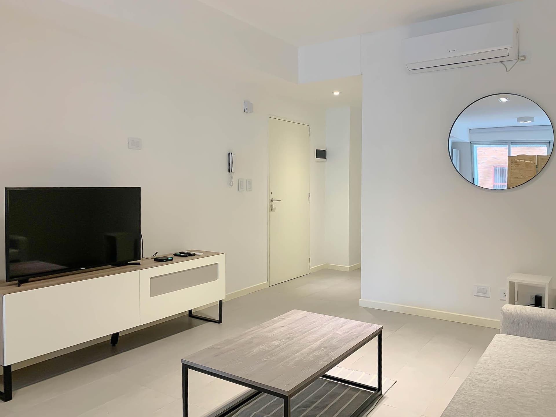 Living con televisor, espejo redondo, aire acondicionado y puerta de entrada.