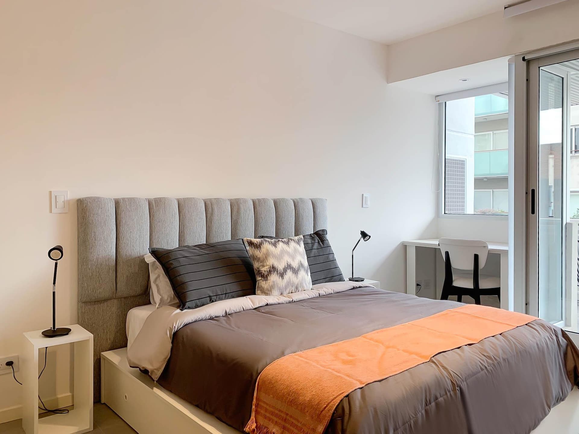 Dormitorio con cama, cabecera gris, lámpara de noche y escritorio.