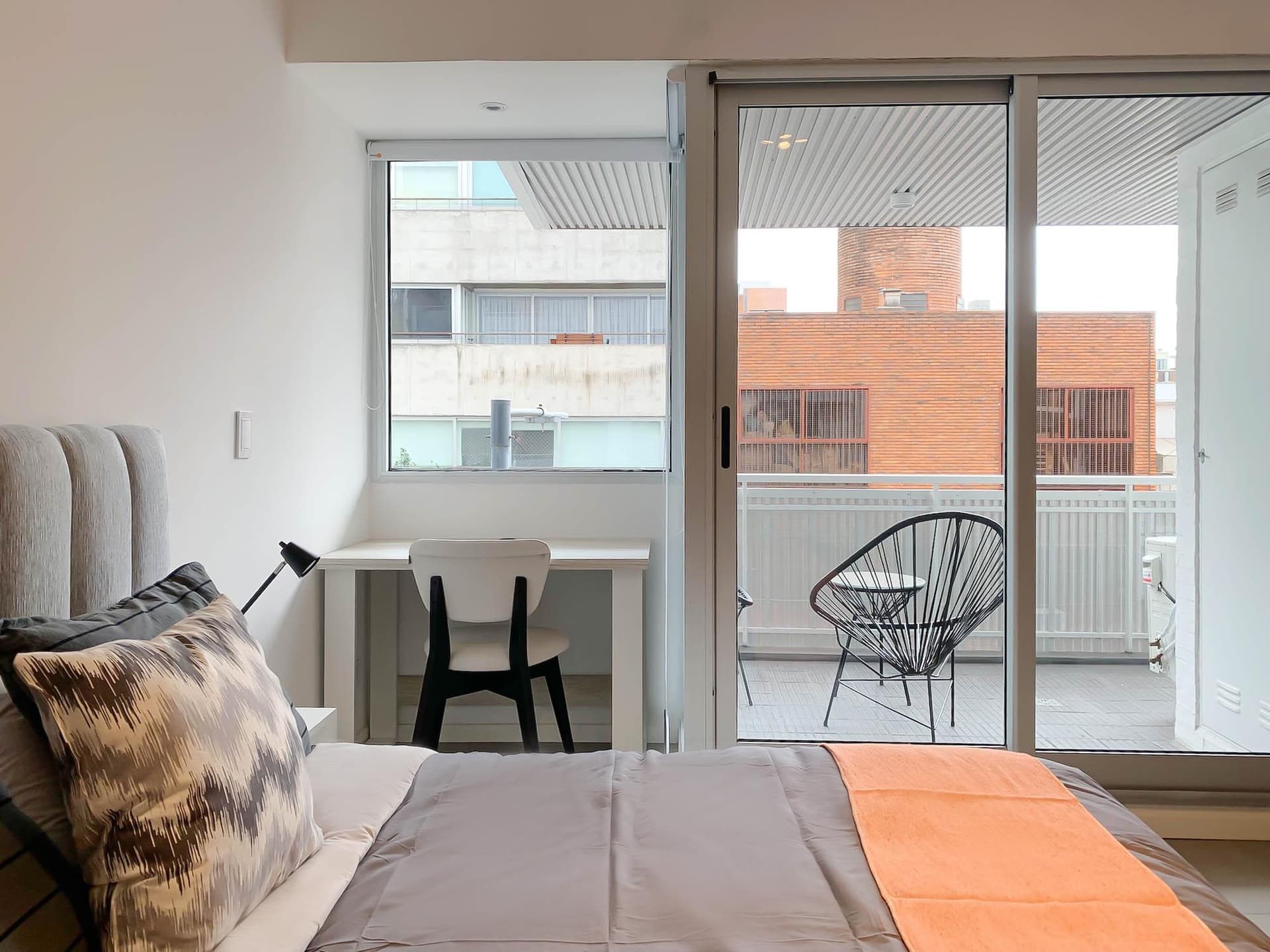 Dormitorio con cama, escritorio blanco y balcón con silla.