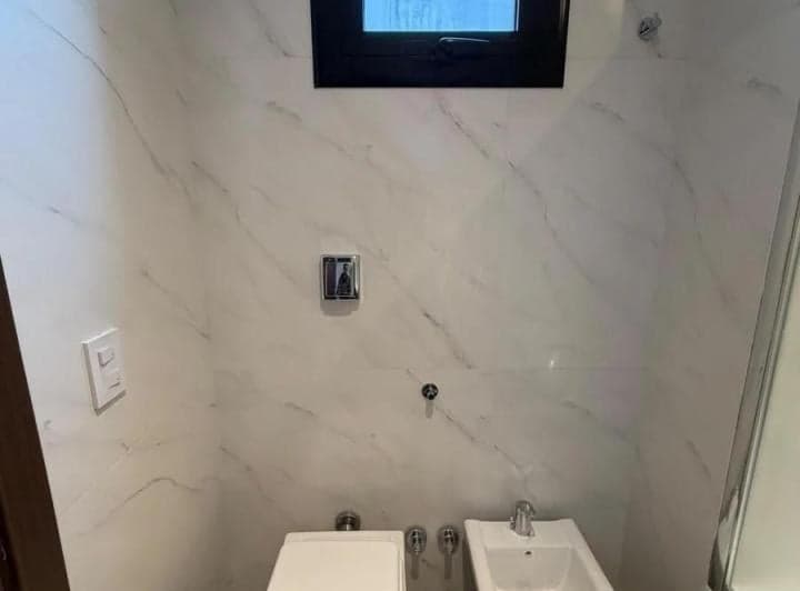 Baño con azulejos de mármol, inodoro, bidet y ventana pequeña.