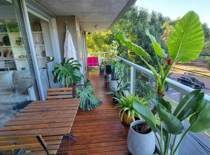 Balcón con plantas, mesa y sillas, con vista al exterior y baranda de vidrio.