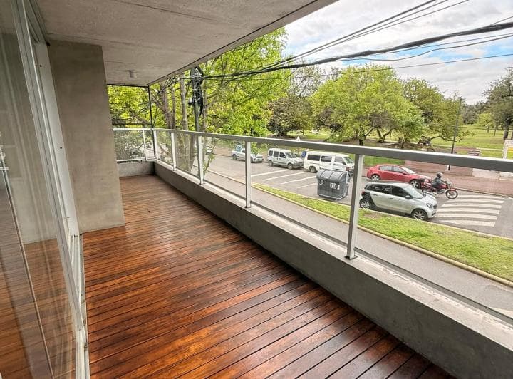 Balcón vacío con piso de madera y baranda de vidrio, con vista a la calle.