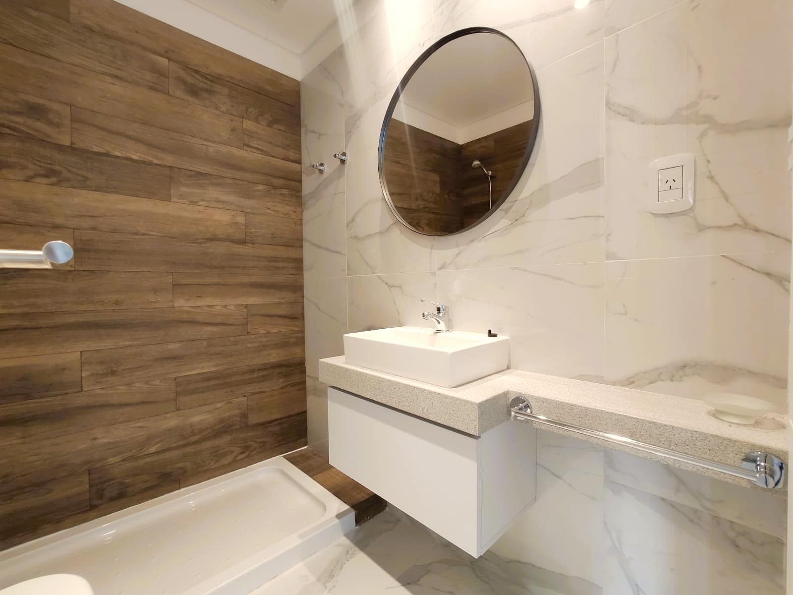 Baño con vanitory, espejo redondo y ducha con azulejos de madera.