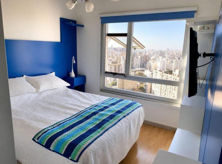 Dormitorio principal con cama doble, cabecera azul y ventana con vista a la ciudad.