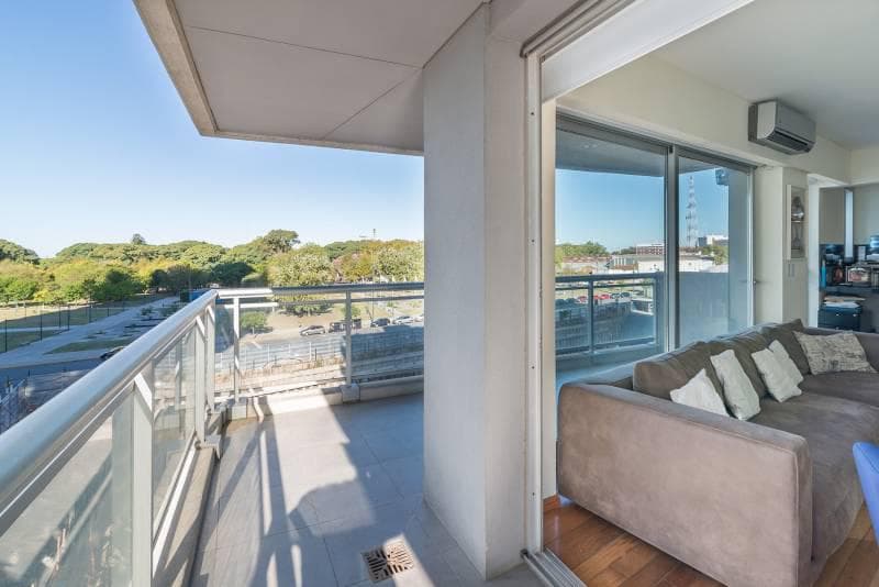 Balcón con baranda de vidrio, vista a un parque y puerta al living.
