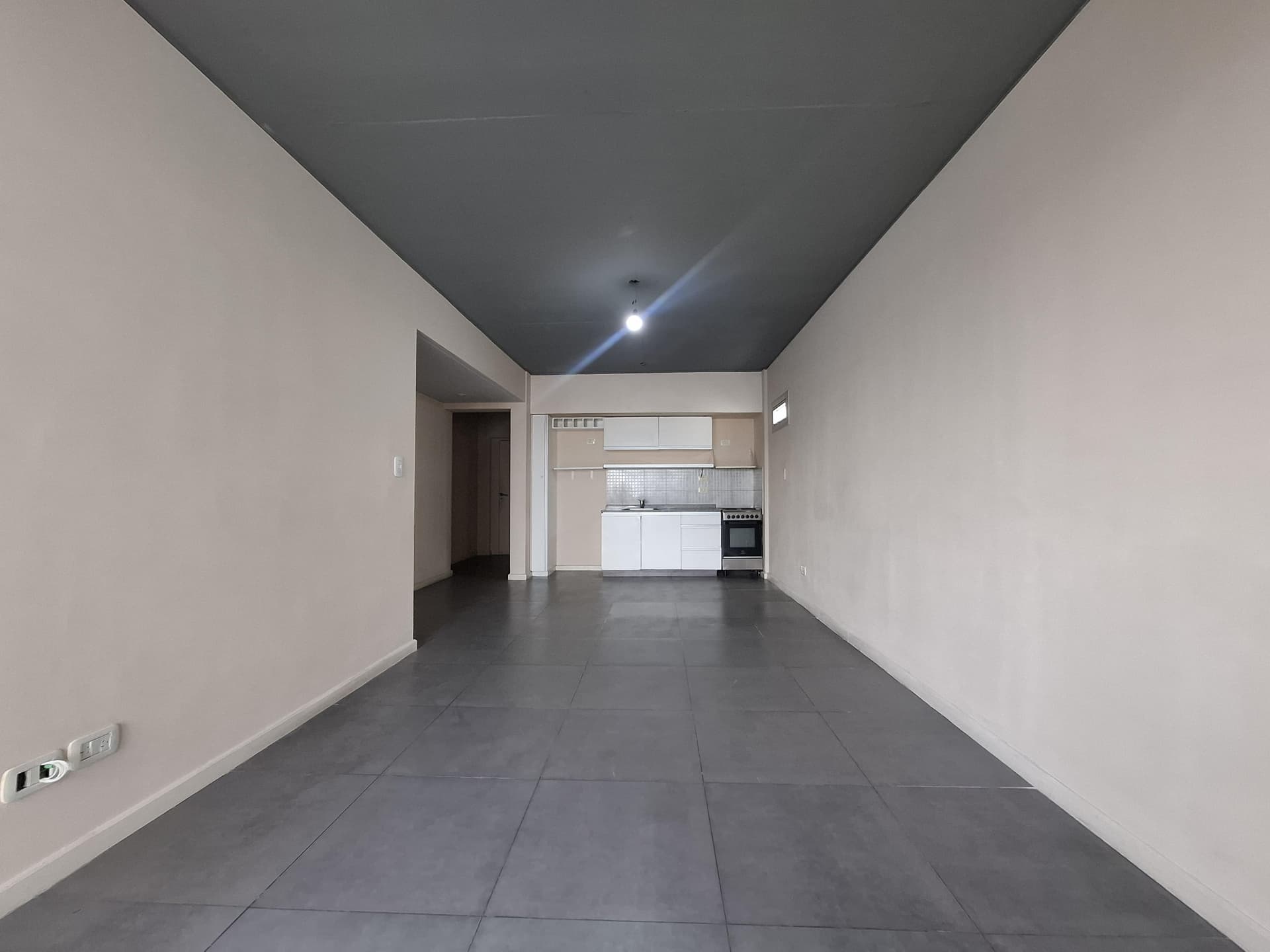 Living de departamento vacío con cocina integrada y techo oscuro.