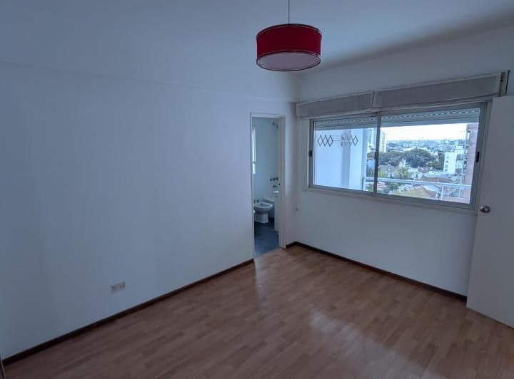 Dormitorio vacío con piso de madera, ventana y vista parcial al baño.