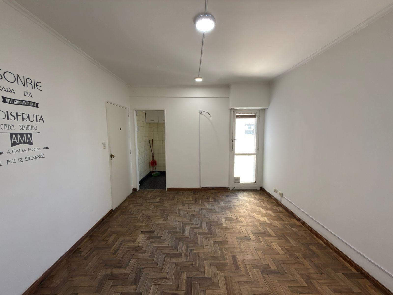 Living vacío con piso de parquet, acceso a cocina y salida al exterior.