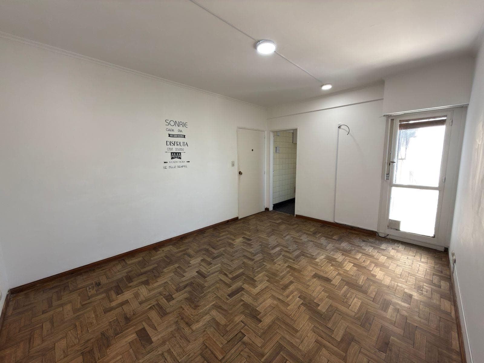 Living vacío con piso de parquet, paredes blancas y puerta de acceso.