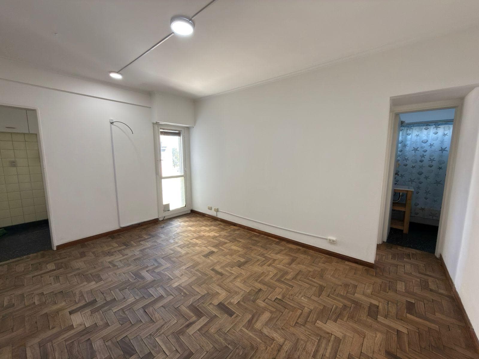 Living vacío con piso de parquet, ventana y puerta a otro ambiente.
