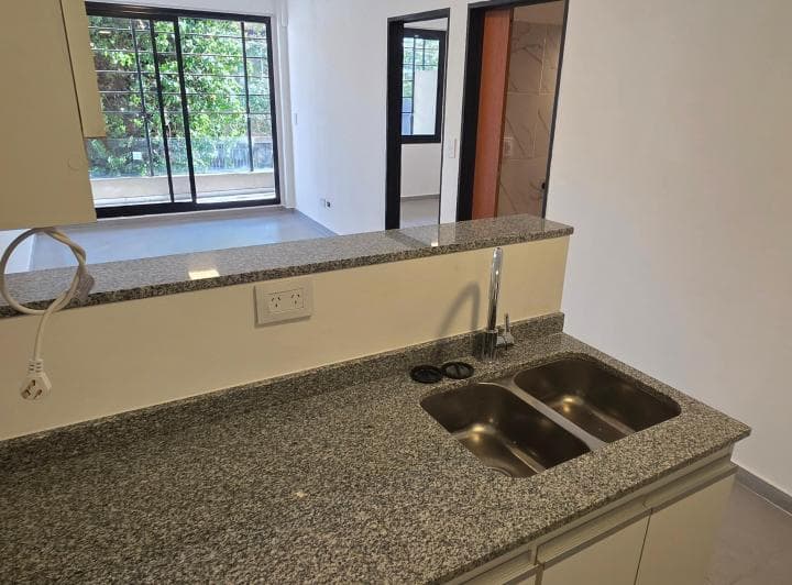 Cocina con bacha, mesada de granito y vista al living comedor vacío con balcón.