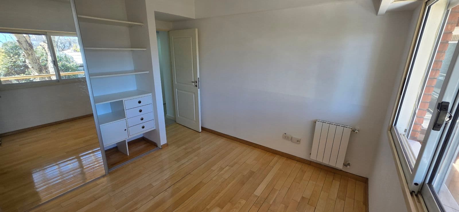 Dormitorio vacío con piso de madera, placard con espejo y ventana.