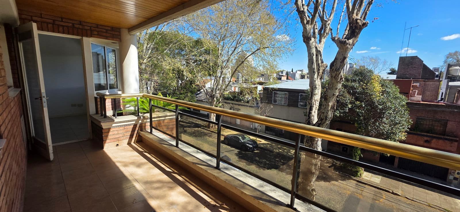 Balcón con baranda de vidrio, salida desde el living y árboles en la calle.