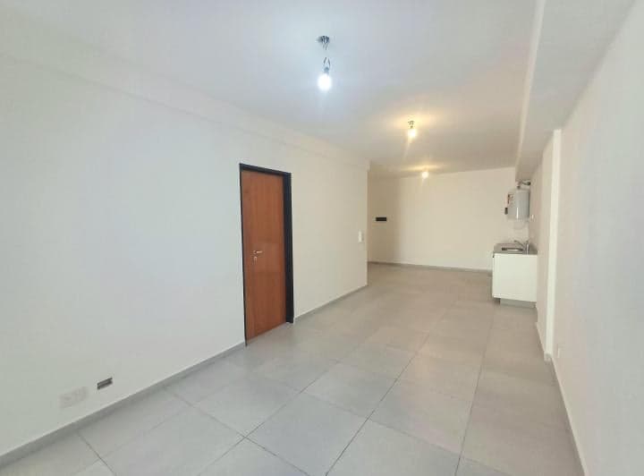 Amplio ambiente vacío con piso de baldosas, puerta de madera y cocina integrada al fondo.